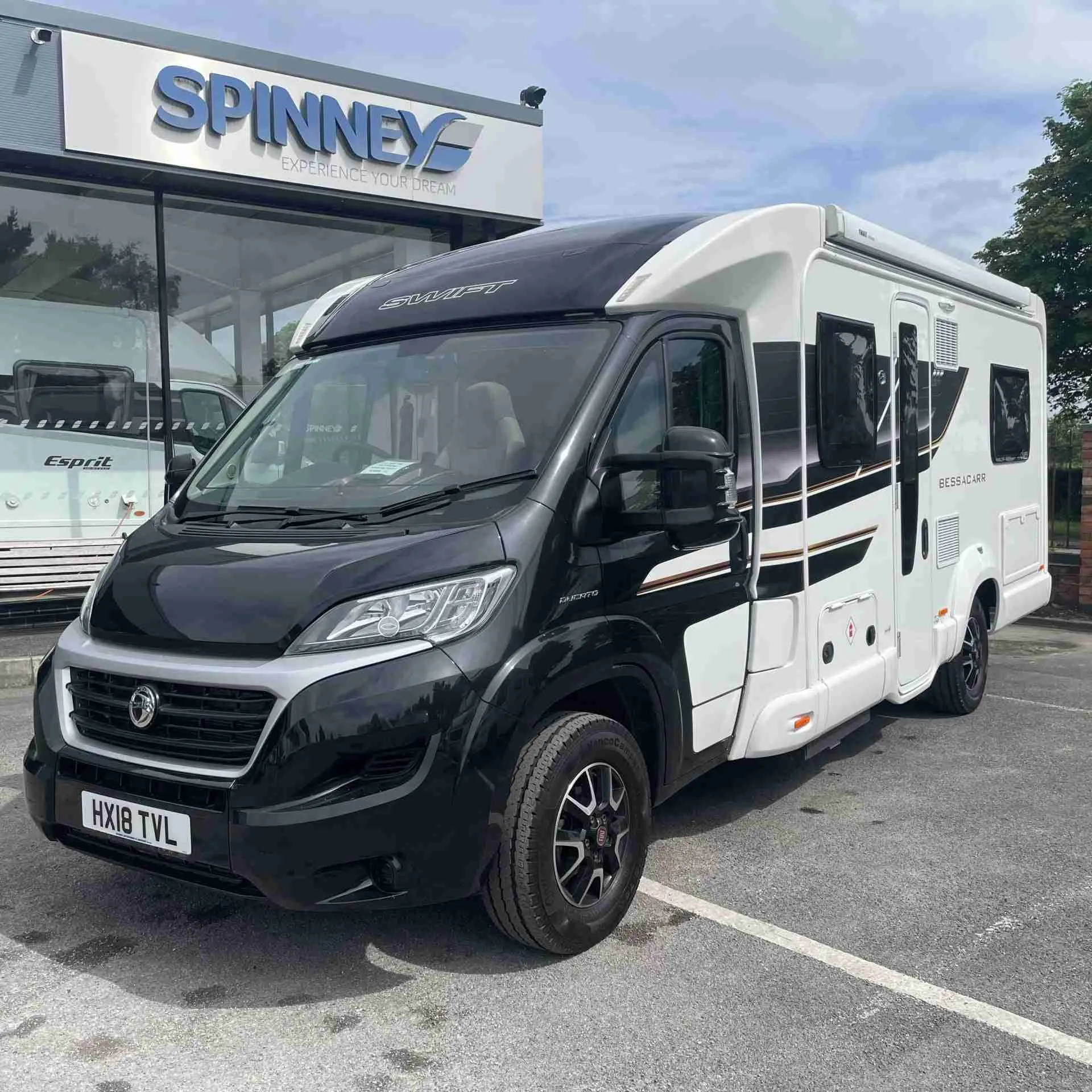 A modern Swift 6 berth white motorhome with black trim parked in front of a dealership named "Spinney" under a clear sky.